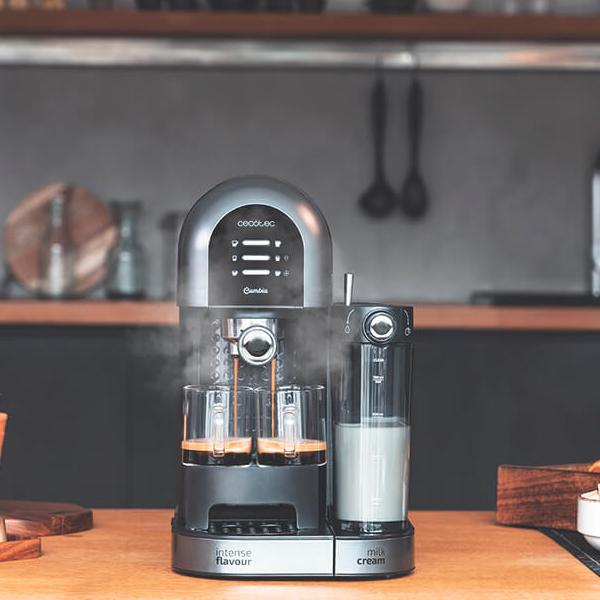 Macchina da caffè con vapore, due bicchieri di caffè e un contenitore di latte su un tavolo di legno.