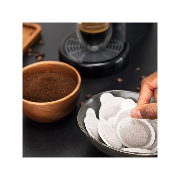 Osoba wyjmuje kapsułki kawy z miski, z mieloną kawą i ekspresem w tle.