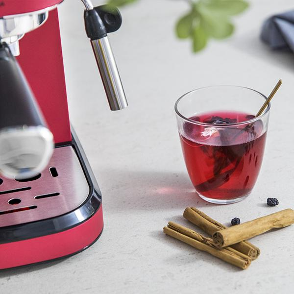 Czerwony ekspres do kawy ze szklanką czerwonej herbaty i laskami cynamonu. Widoczne są liście i jagody.