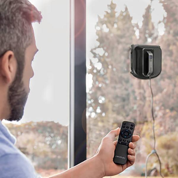 Mann benutzt Fernbedienung, um Fenster mit Roboterreiniger zu putzen. Bäume im Hintergrund.