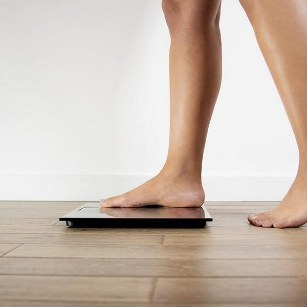 Stopy osoby na wadze, z drewnianą podłogą i białą ścianą w tle.