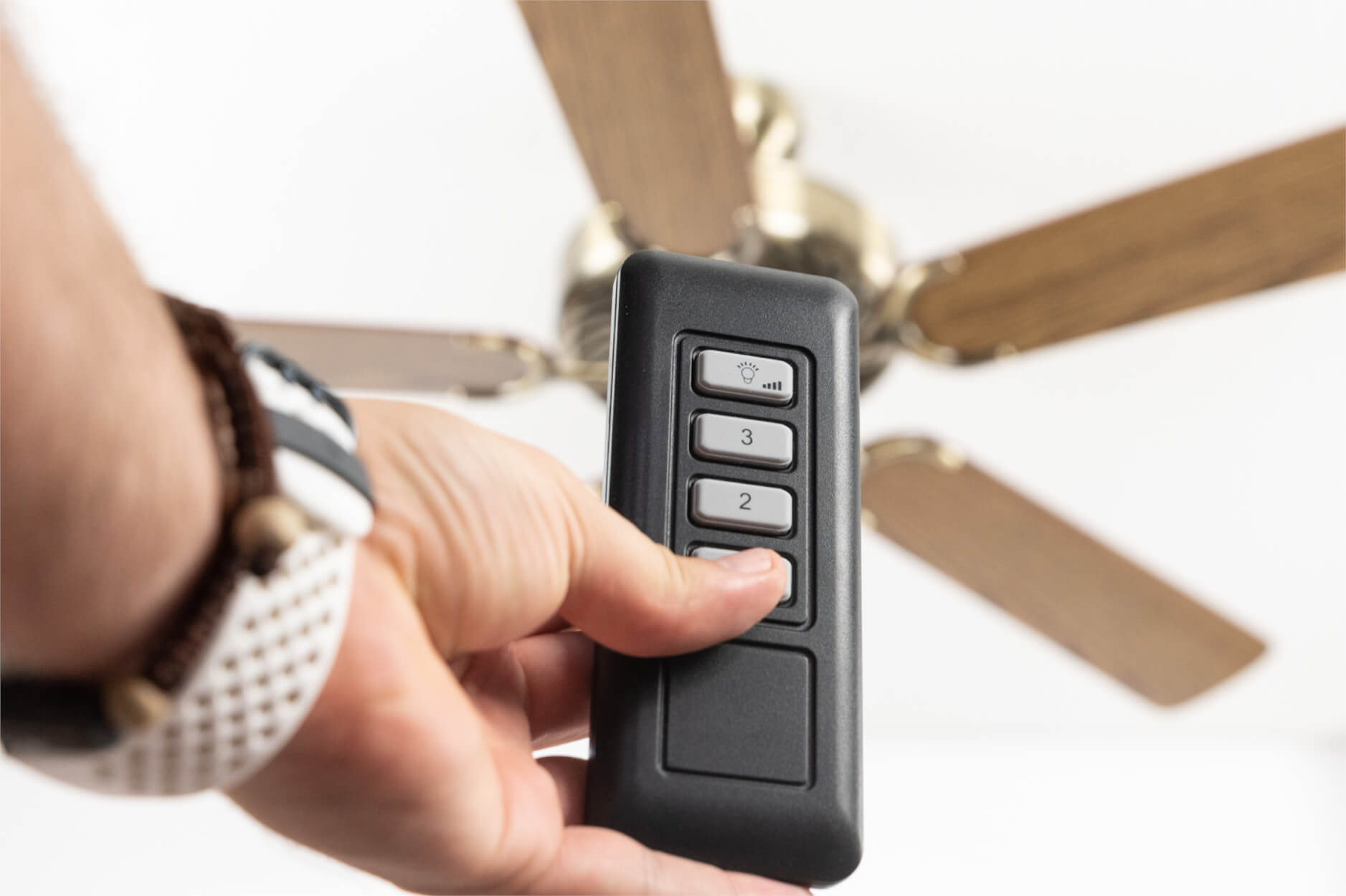 Eine Hand, die eine Fernbedienung hält und auf einen Deckenventilator zeigt. Der Ventilator ist teilweise sichtbar. Weißer Hintergrund.
