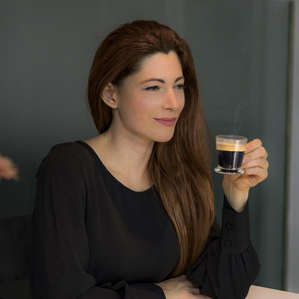 Kobieta trzymająca małą szklankę kawy. Ma długie brązowe włosy i nosi czarną koszulę.