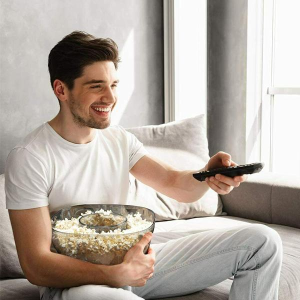 Un uomo su un divano tiene in mano una ciotola di popcorn e un telecomando, sorridendo.