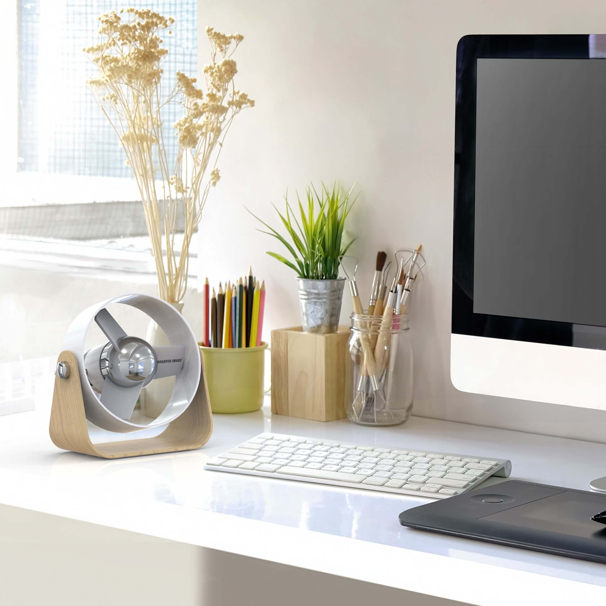 Ein weißer Schreibtisch mit Ventilator, Bleistiften, Pinseln und einem Computer.