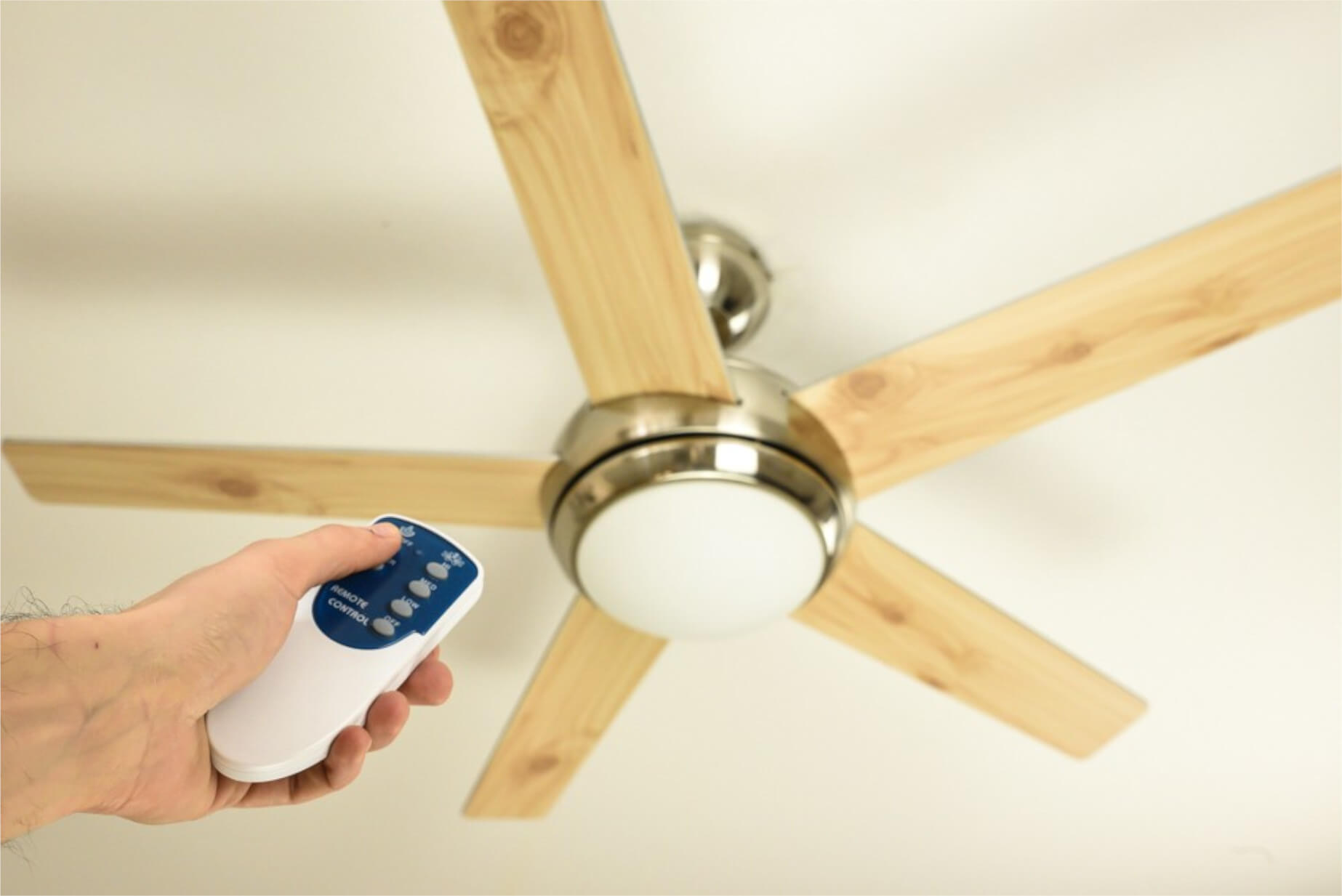 Eine Hand, die eine weiße Fernbedienung hält, die einen Deckenventilator mit Holzflügeln steuert.