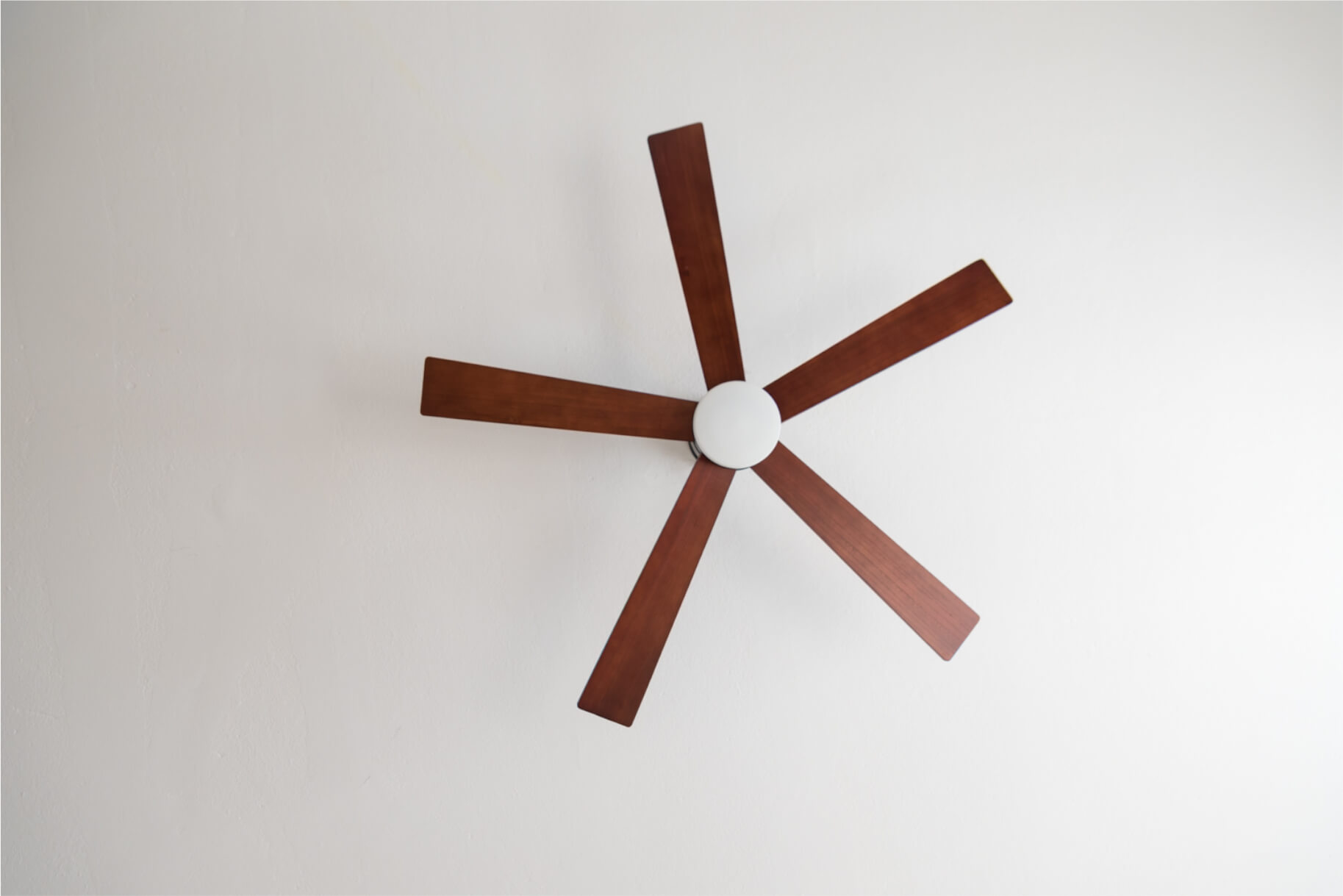 Ein Deckenventilator mit fünf braunen Flügeln und einem weißen Zentrum von unten vor weißer Decke gesehen.