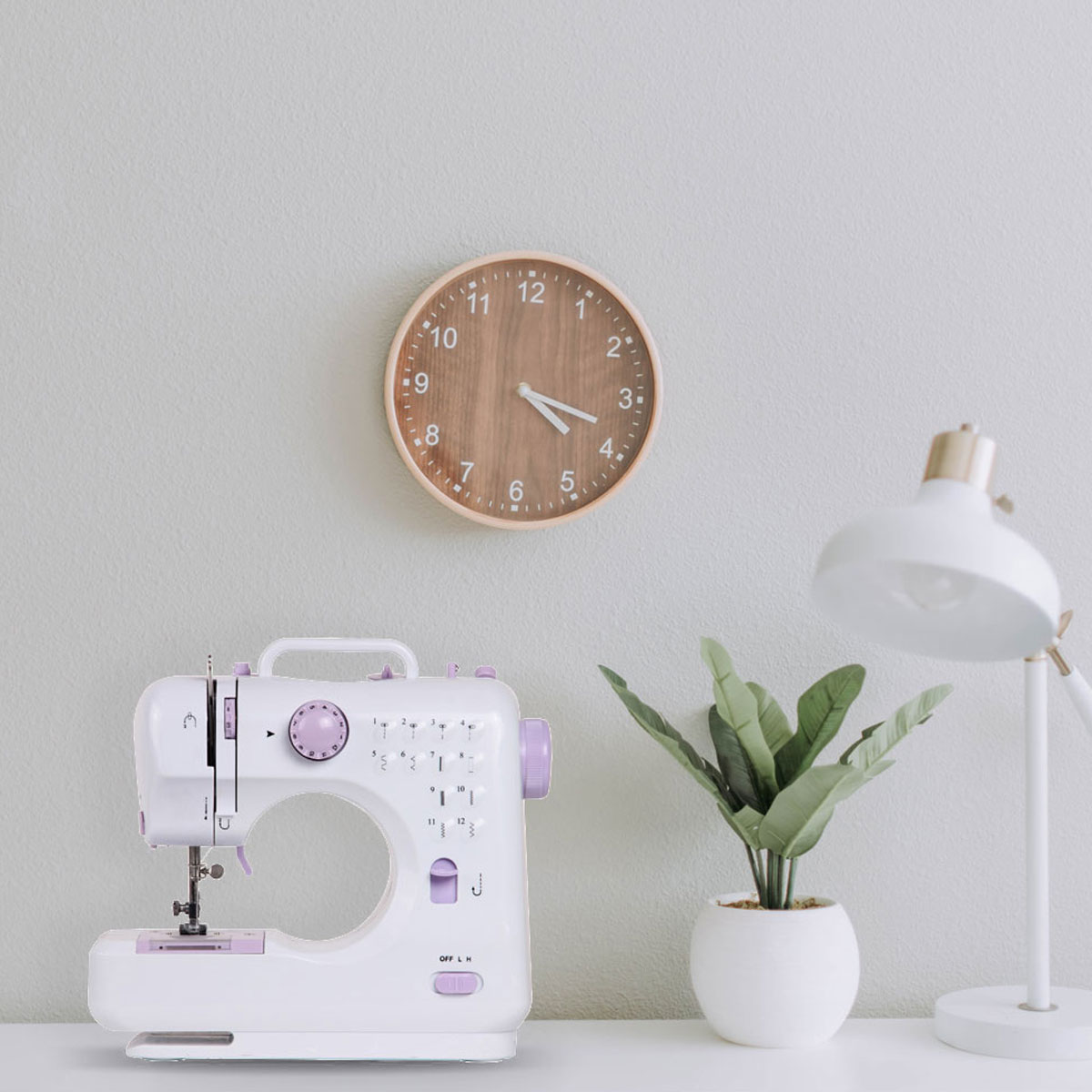 Maszyna do szycia, zegar, roślina i lampa na białym stole na tle szarej ściany. To ustawienie domowego biura.