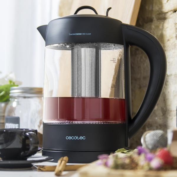 Hervidor negro con cuerpo de vidrio. Té rojo en el interior. Una taza y algunas flores están en el fondo.
