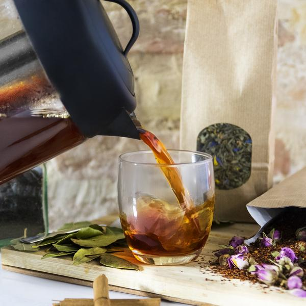 Té que se vierte de una tetera negra en un vaso. Bolsas de papel marrón y especias en el fondo.