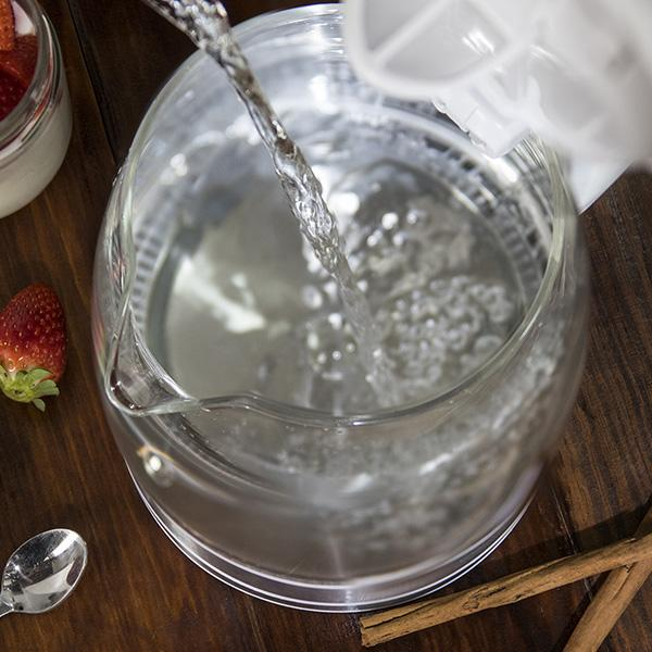 Wasser wird in einen transparenten Glaskessel gegossen, Erdbeeren und Löffel in der Nähe.