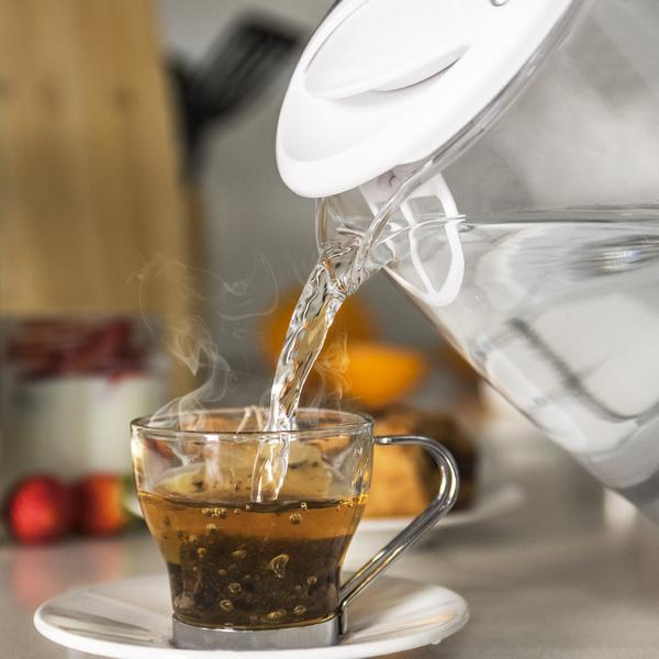Klares Glas gießt heißes Wasser in eine Glastasse mit Teebeutel. Weiße Untertasse. Unscharfer Hintergrund.