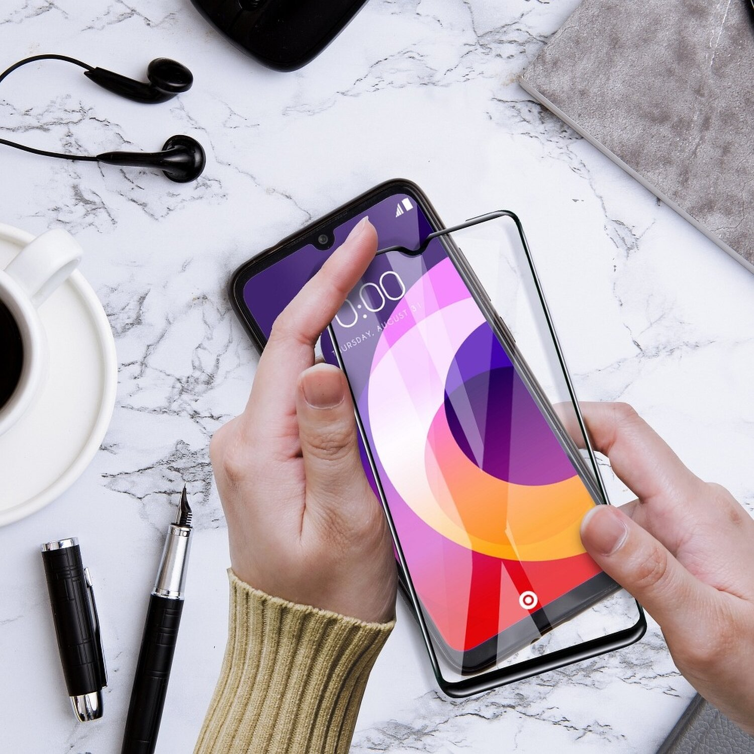 Hände, die ein Telefon mit Displayschutzfolie halten. Kopfhörer, Stift und Tasse auf Marmoroberfläche.