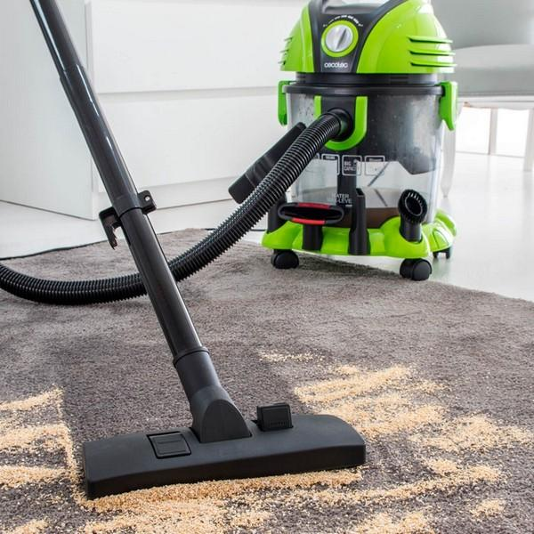 A vacuum cleaner with a green and black body is cleaning a carpet, with sawdust.