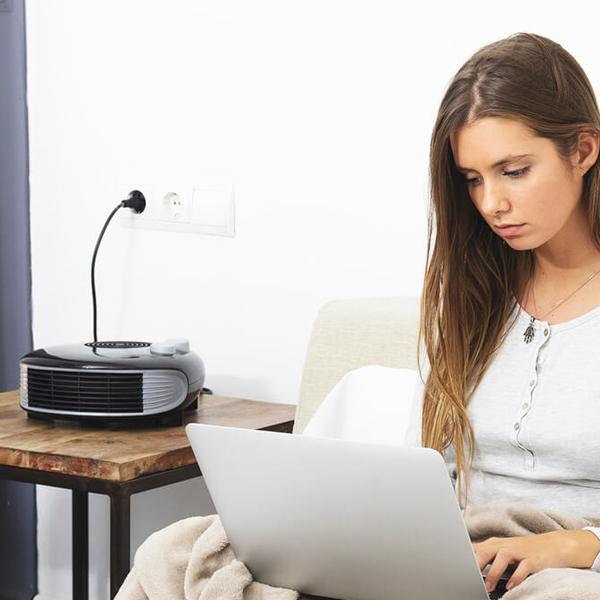 Een vrouw die een laptop gebruikt op een bed. Een kleine kachel staat op een tafel. De muren zijn wit.