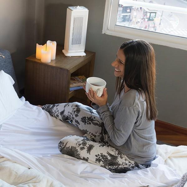 Kobieta siedzi na łóżku, uśmiecha się, trzyma biały kubek, ze świecami i grzejnikiem.