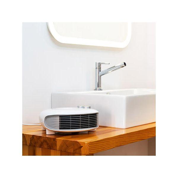 Biały grzejnik elektryczny stoi na drewnianej powierzchni przed białą umywalką i lustrem.