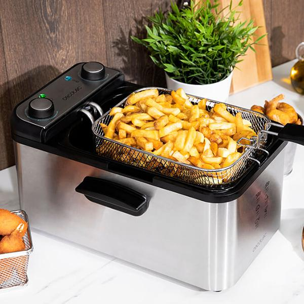 Eine Edelstahl-Fritteuse mit Pommes im Korb; neben Schneidebrett und Pflanzen.