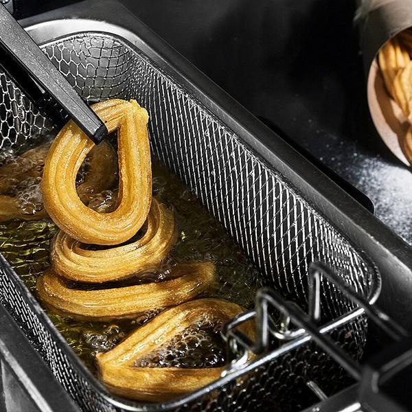 Churros smażone w oleju, trzymane przez szczypce w metalowym koszu.