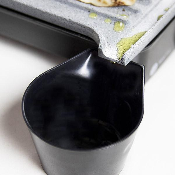 Zbliżenie czarnego pojemnika z szarą kamienną płytą grillową. Olej kapie z płyty.