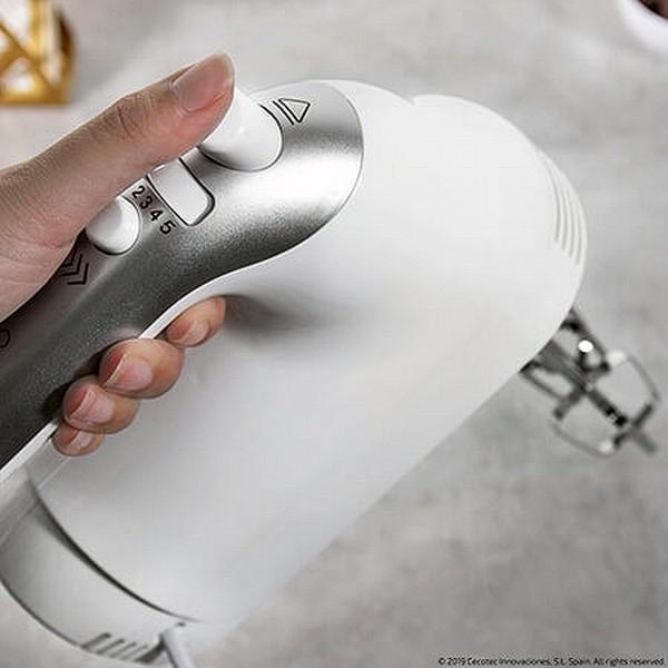 Eine Hand benutzt einen elektrischen Mixer. Der Mixer ist weiß, mit einem silbernen und grauen Bedienfeld.