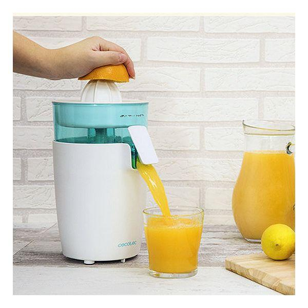 Eine Person presst Orangensaft mit einem weißen und tealfarbenen Entsafter, Glas und Saftkanne im Hintergrund.