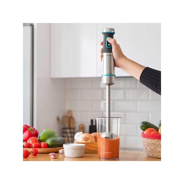 Hand die een zilveren staafmixer vasthoudt in een glas met rode vloeistof en groenten.