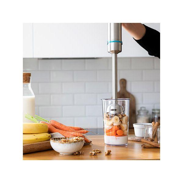 Hand hält einen Mixer und mischt Zutaten. Bananen, Karotten, Milch und Nüsse. Küchenszene.