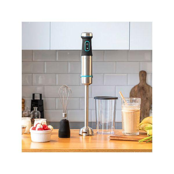 Ein Stabmixer mit Zubehör. Eine Schüssel mit Obst und ein Smoothie-Glas stehen auf der Theke.
