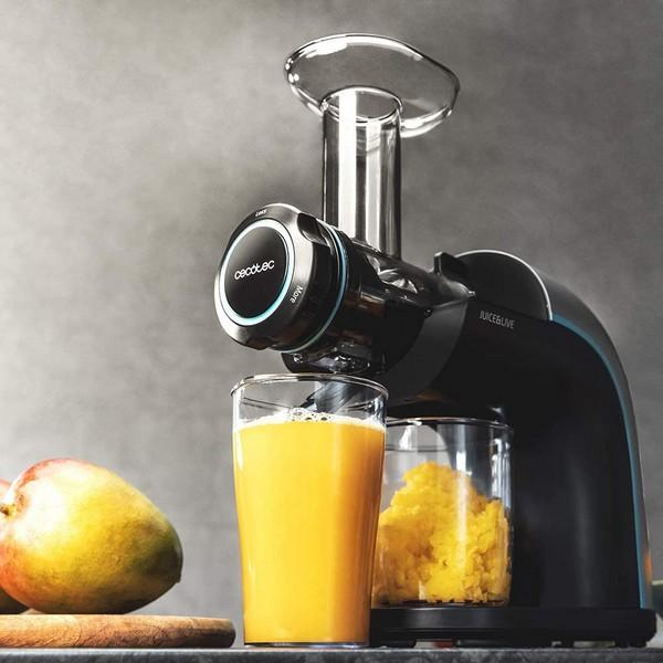 Entsafter mit einem Glas Saft und Fruchtfleisch sowie einer Mango auf einem Holzbrett, grauer Hintergrund.