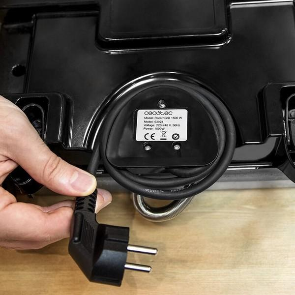 Ręka trzymająca czarny przewód zasilający z wtyczką, podłączony do czarnego urządzenia.