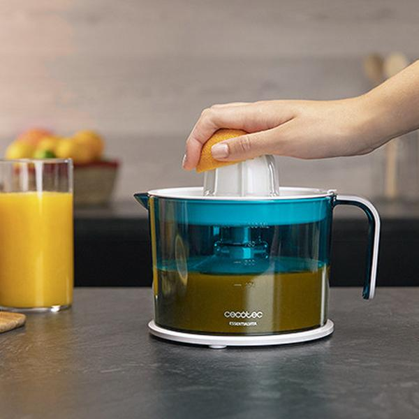 Una mano che sprema metà arancia in uno spremiagrumi. Lo spremiagrumi è verde acqua e bianco, con un bicchiere.