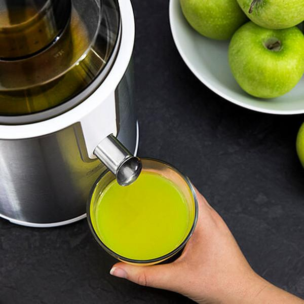 Una exprimidora con un vaso de jugo verde y manzanas verdes en un cuenco en el fondo.