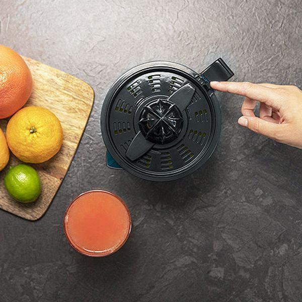 Una centrifuga con una mano che preme un pulsante, accanto a arance, lime e succo in un bicchiere.