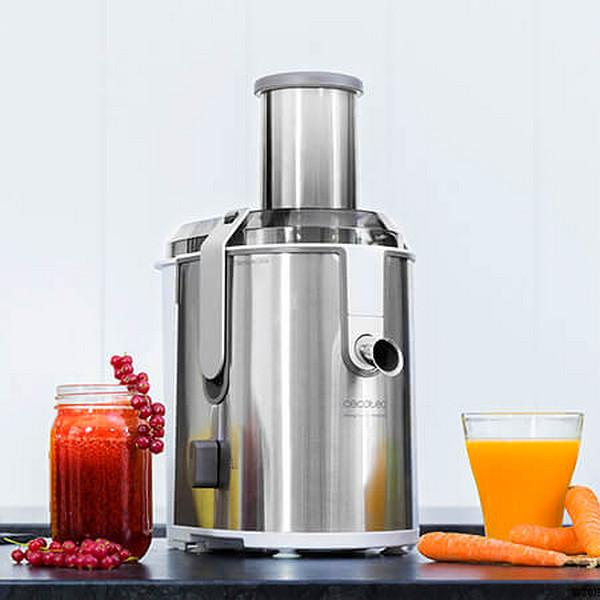 Exprimidor plateado con un vaso de zumo de naranja y un tarro de zumo rojo con zanahorias y bayas al lado.