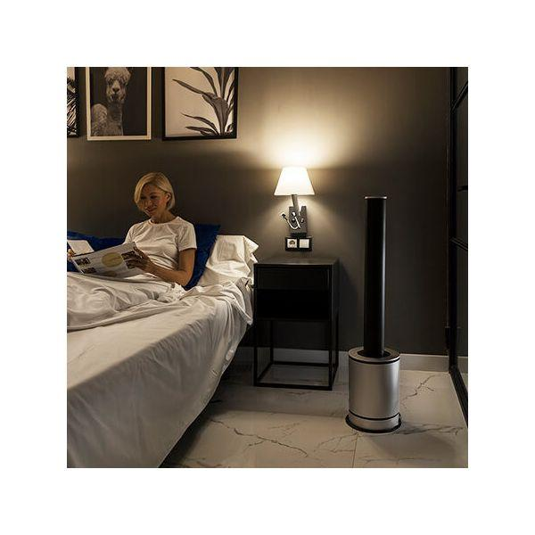 Donna che legge a letto, lampada a muro, ventilatore a terra, quadri a muro.