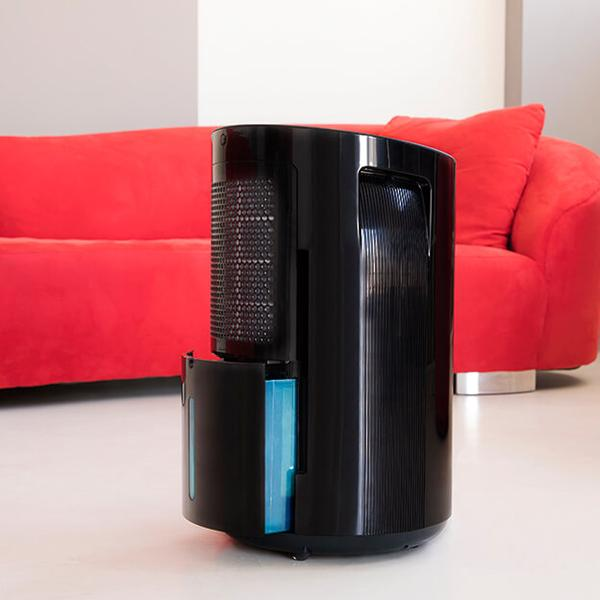 Czarny oczyszczacz powietrza z niebieskim zbiornikiem na wodę, na białej podłodze, z czerwoną sofą w tle.