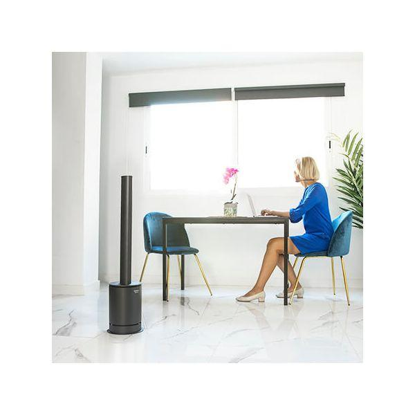 Una donna che lavora a una scrivania con un laptop. Un ventilatore a torre nero è sul pavimento.