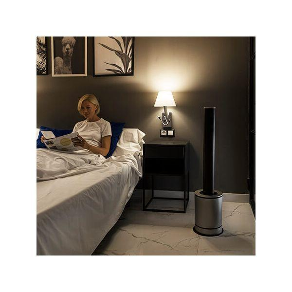 Donna che legge a letto con una lampada sul comodino. Un ventilatore cilindrico alto vicino al letto.
