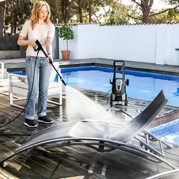 Mujer usa hidrolavadora en tumbona gris junto a piscina. Lavadora negra al fondo.