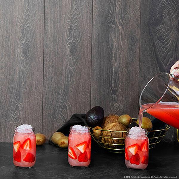 Tres frascos de bebida rosa con fresas y hielo, con otras frutas y una persona vertiendo.