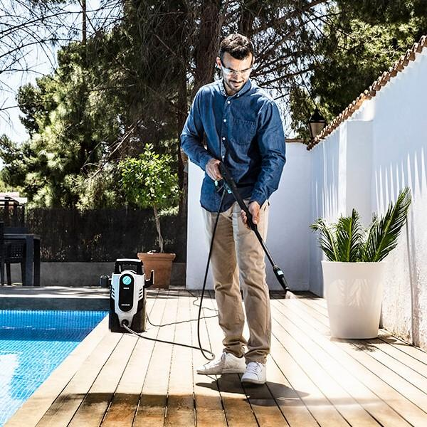Hombre usando una hidrolavadora en una terraza de madera. El fondo muestra una casa y árboles.