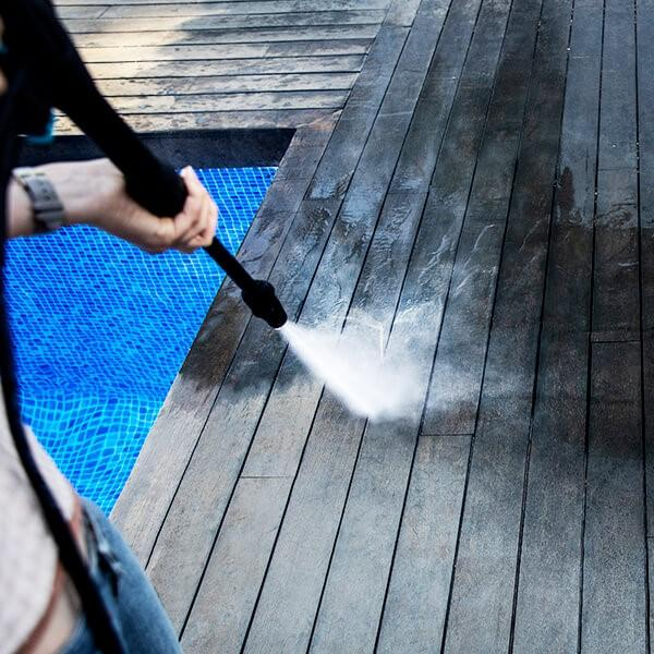 Persona limpiando terraza de madera con hidrolavadora. Piscina azul al fondo.