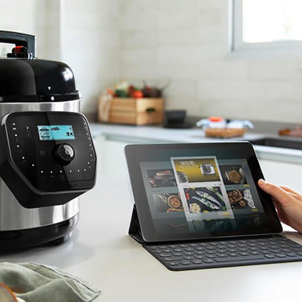 Un electrodoméstico negro y plateado con una pantalla digital. Una tableta muestra recetas cerca. Una mano la está tocando.