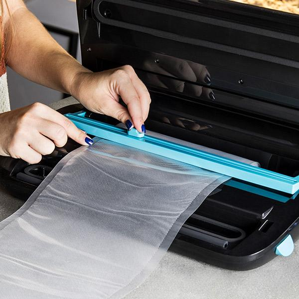 Ręce kobiety zamykają worek. Zgrzewarka próżniowa, czarna i turkusowa. Worek jest przezroczysty.