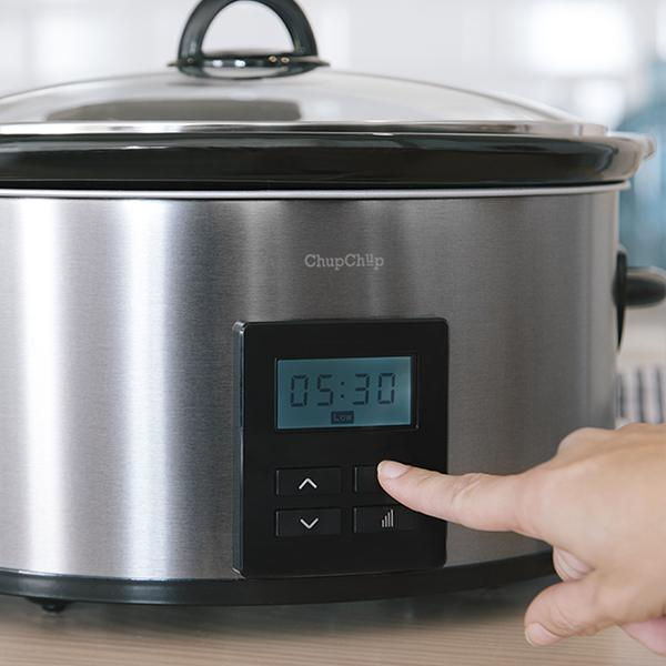 Una olla de cocción lenta de acero inoxidable con un panel de control negro muestra la hora. Una mano pulsa un botón.