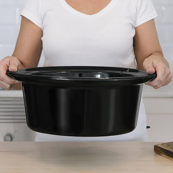 Una persona sosteniendo una olla de cocción lenta negra. La olla está vacía sobre una mesa de madera.
