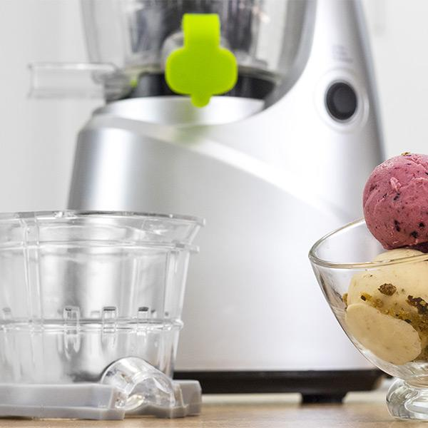 Máquina plateada para hacer helado con acento verde, partes transparentes y dos bolas de helado en un bol de vidrio.