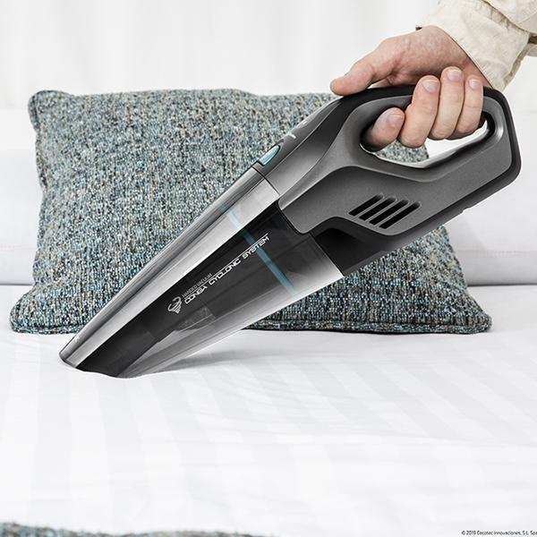 Aspiradora de mano en una cama. La aspiradora es gris y negra. Una almohada está detrás.