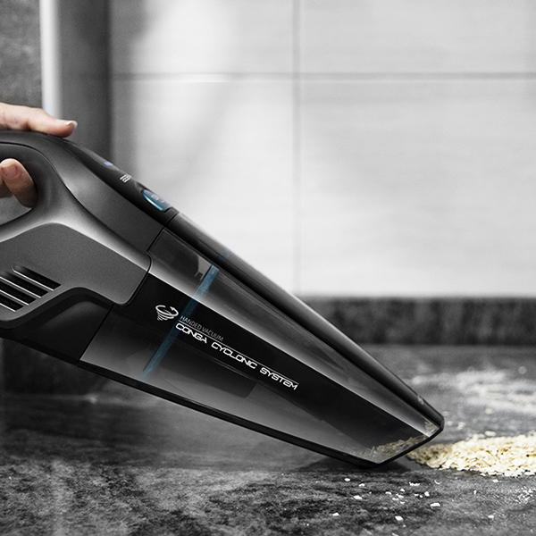 Aspirateur à main noir nettoyant une surface. Des débris beiges sont visibles sur la surface.